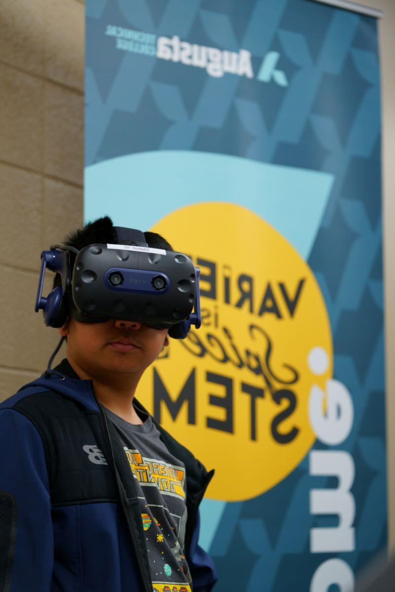 An African American male stands solemnly wearing VR equipment in front of an Augusta Technical College VARIETY is Spice STEM banner.